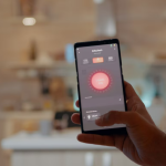 celular na mão de uma pessoa em uma cozinha com luz indireta aconchegante, na tela, mostra a pessoa está controlando a temperatura do ambiente por meio do celular, ilustrando o tema do blog que é casas inteligentes